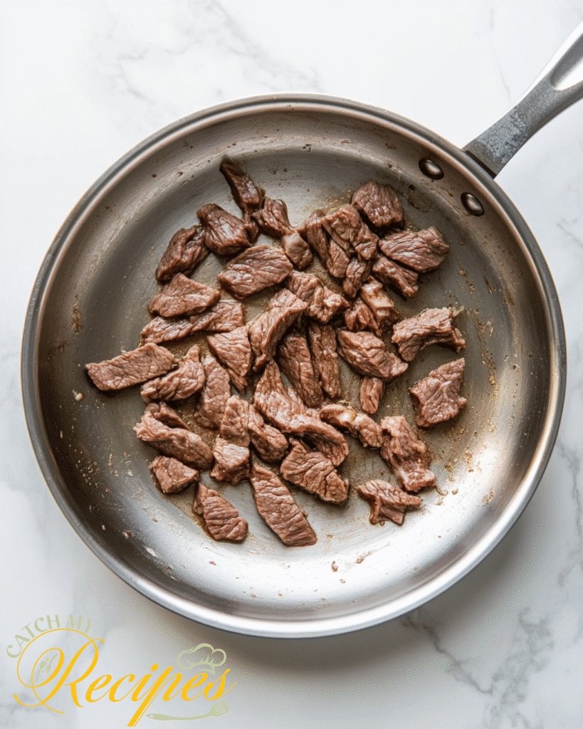 Beef Stroganoff Recipe Without Mushrooms 4 Sautéed beef strips in a stainless steel frying pan on a white marble surface.